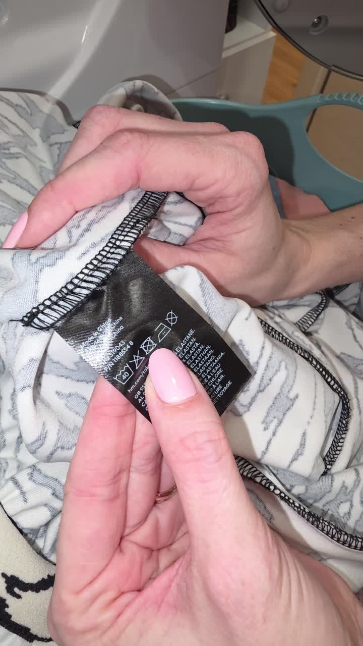 Vertical close-up of hands reading a clothing care label with a do not tumble dry symbol at home in a laundry room, helping prevent shrinkage and heat damage