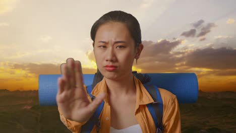 close up of asian female hiker with mountaineering backpack looking at camera and making stop hand sign while standing on the top of mountain during sunset time