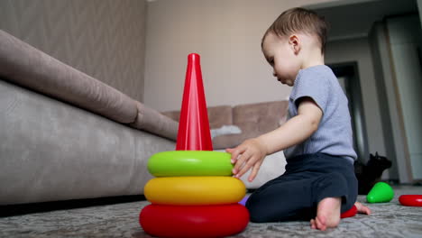 cute caucasian kid playing with toy pyramid. child plays on the floor at home. black cat at backdrop.