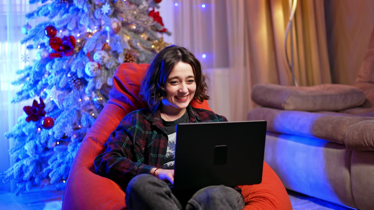 Happy person using laptop cozy. A young person sits comfortably in a bean bag, using a laptop, surrounded by festive decorations and a Christmas tree