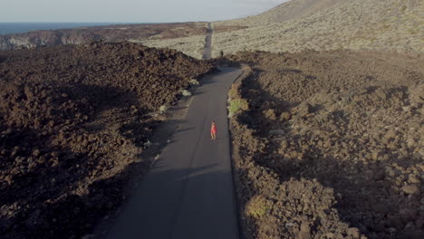 mujer enérgica vestida de rojo caminando por una carretera desierta en un valle entre montañas y costa rocosa en una tarde soleada, isla de hierro, canario - vista de drones
