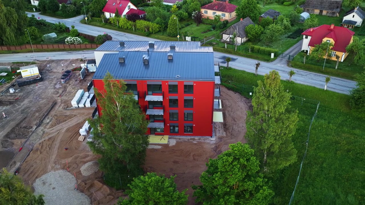 Moderne rød leilighetsbygning omgitt av forstadshus, hager og trær ved siden av en byggeplass