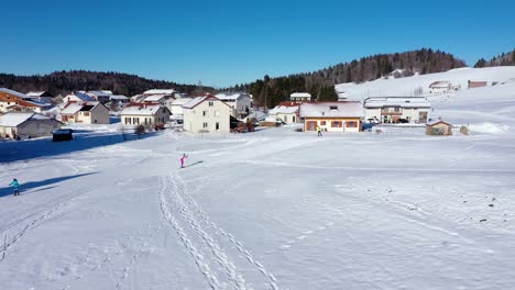 Toma-Aérea-De-Un-Atleta-De-Biatlón-Con-Traje-De-Esquí-Rosa-Esquiando-Cuesta-Arriba-En-Una-Pista-Nevada-Junto-A-Casas-De-Apartamentos-En-Jura,lajoux,francia