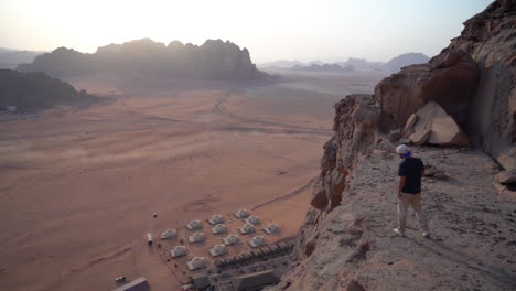 Hombre-Caminando-En-Un-Acantilado-Sobre-El-Campamento-Beduino-Y-El-Paisaje-Desértico-De-Wadi-Rum,-Jordan-Con-Horizonte-Brumoso,-Cámara-Lenta