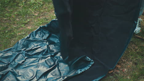 close up of dark blue tent fabric unfolding on grassy forest ground, showing wrinkled texture and motion as someone opens it during outdoor camping activity in natural environment