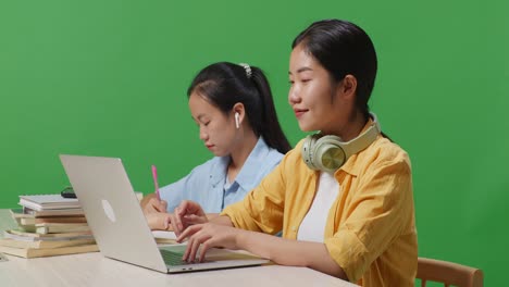 close up of asian woman student typing on a laptop and talking on smartphone while sitting with her friend studying on a table in the green screen background classroom