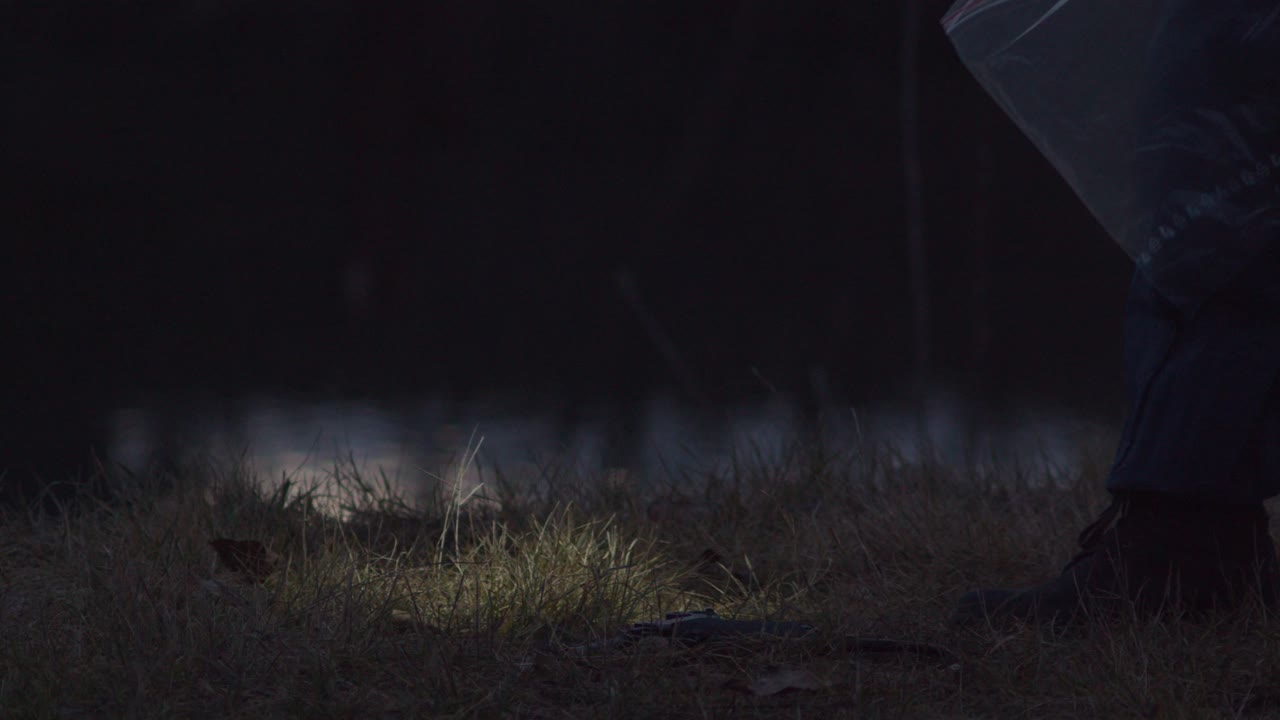 손전등을 든 형사가 땅에서 총을 발견하고 밤에 범죄 현장 증거를 찾았습니다