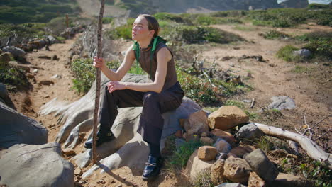 woman resting rocky path with hiking stick in sunlight. fashion model posing