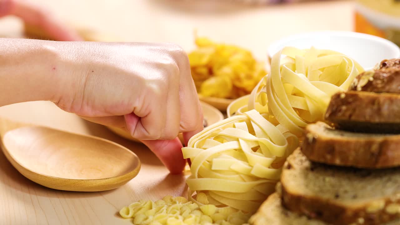 Hands organize grains, pasta, and bread on a wooden table under bright lighting, creating a warm and inviting atmosphere