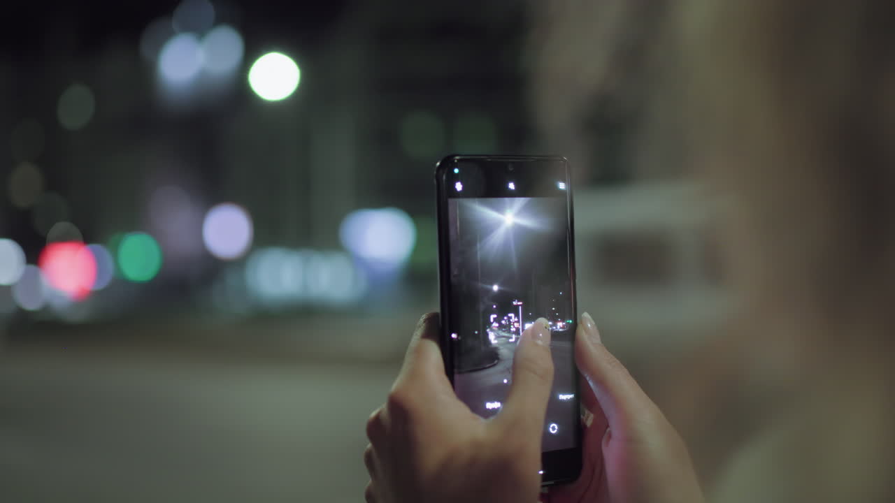Close up of woman using smartphone camera to take photo of illuminated street at night, fingers adjusting focus on screen, colorful city lights in background