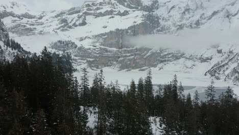 Aerial-of-green-forest-in-a-snow-covered-landscape-with-a-frozen-lake-and-mountains-in-the-background