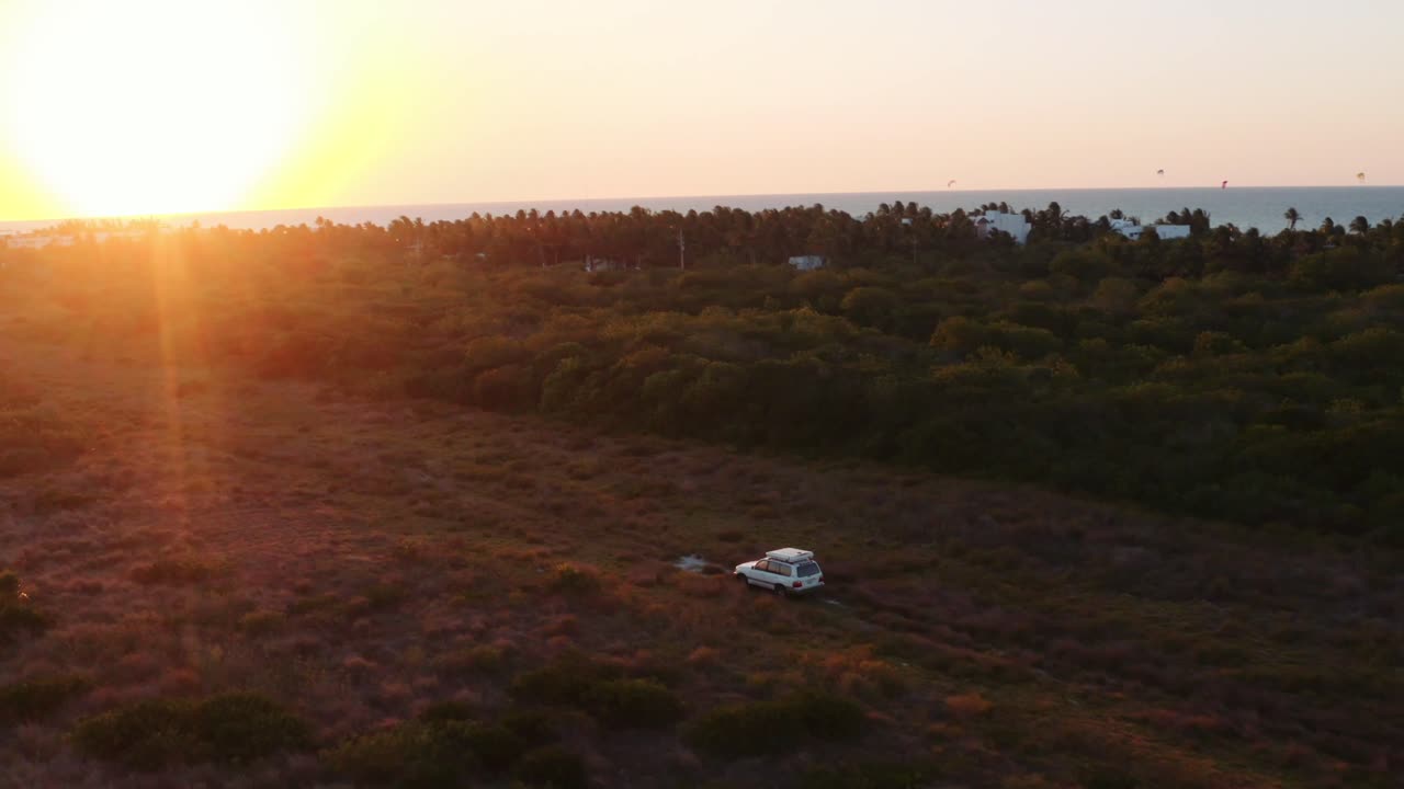 toma aérea amplia y dinámica de todoterreno con una carpa en la azotea que recorre un campo áspero hacia la puesta de sol