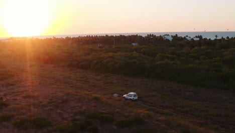 Dynamische-Breite-Luftaufnahme-Eines-Geländewagens-Mit-Einem-Dachzelt,-Das-Durch-Ein-Raues-Feld-In-Richtung-Sonnenuntergang-Fährt