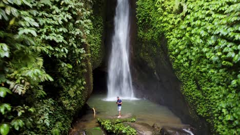 aerial 4k: young tourist male standing at leke leke waterfall, tropical paradise hidden in a lush green moss covered canyon in jungles of bali