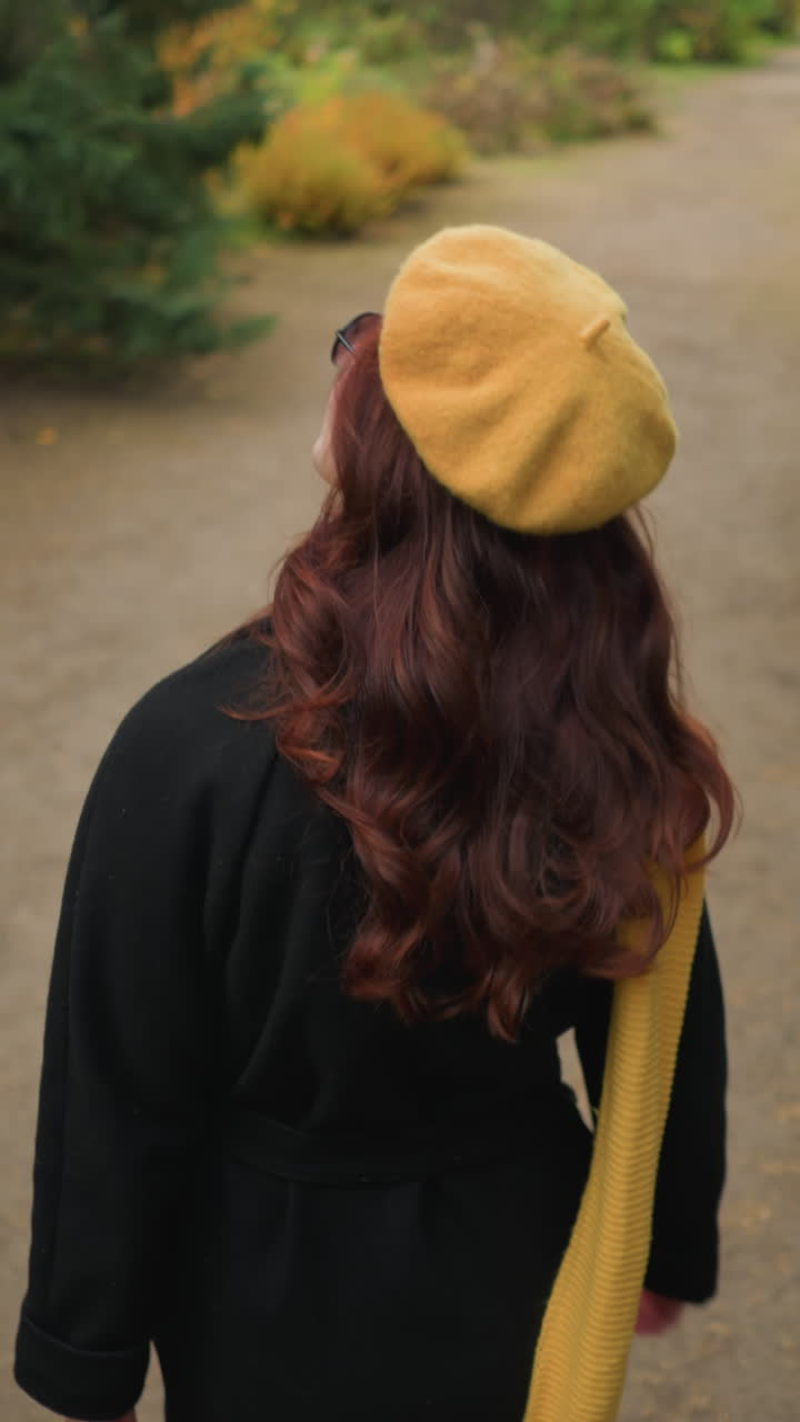 Vista trasera de una dama con boina amarilla y bufanda, ajustándose el cabello mientras pasea por un jardín vibrante lleno de colores otoñales.