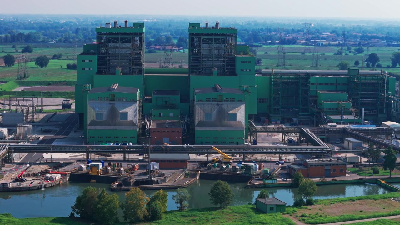 Aerial perspective of a large, green industrial power plant complex with a chimney and surrounding infrastructure next to a river canal