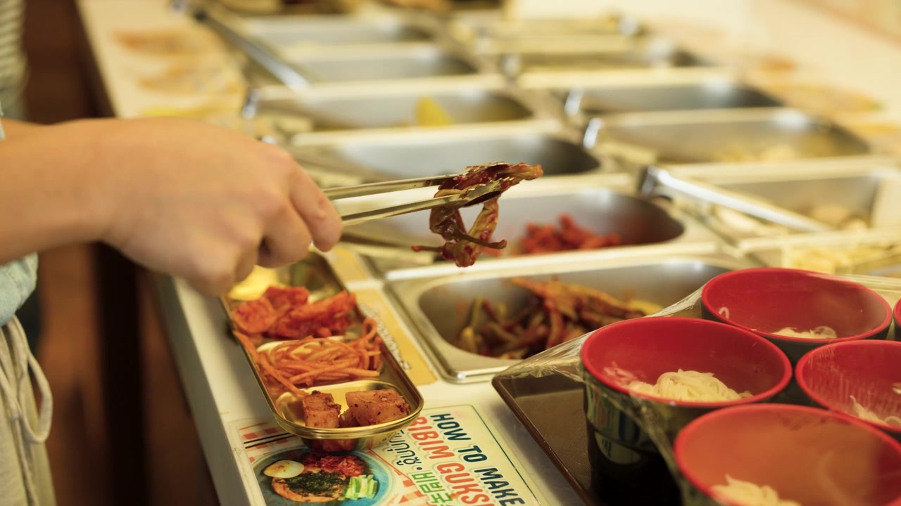 Primo piano di mani che usano pinze di metallo per servire vari contorni banchan al ristorante