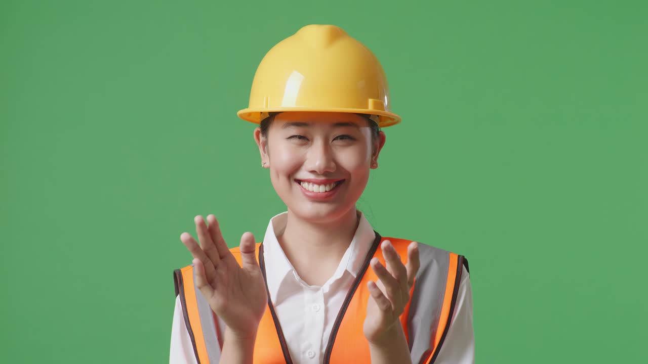 안전 헬을 입은 아시아 여성 엔지니어가 녹색 스크린 배경 스튜디오에 서서 웃고 손을 치고 있습니다.