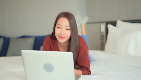 asian woman checking bitcoin price on laptop, suddenly showing a winning expression and yes gesture while lying down on bed