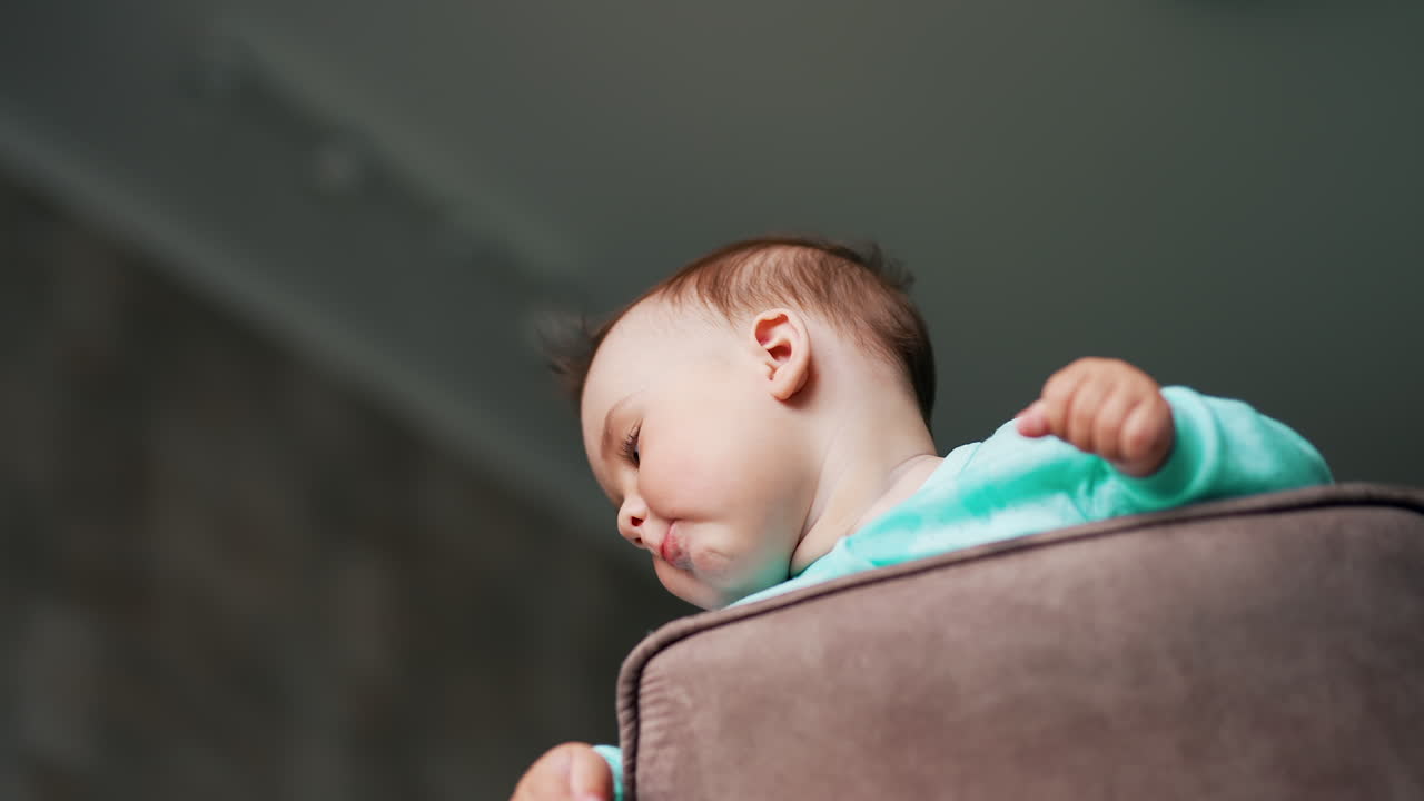 Funny boy with sticking out hair standing on a chair. Kid holding both hands on a chair back and turning his head away from camera. Low angle view. Blurred backdrop.
