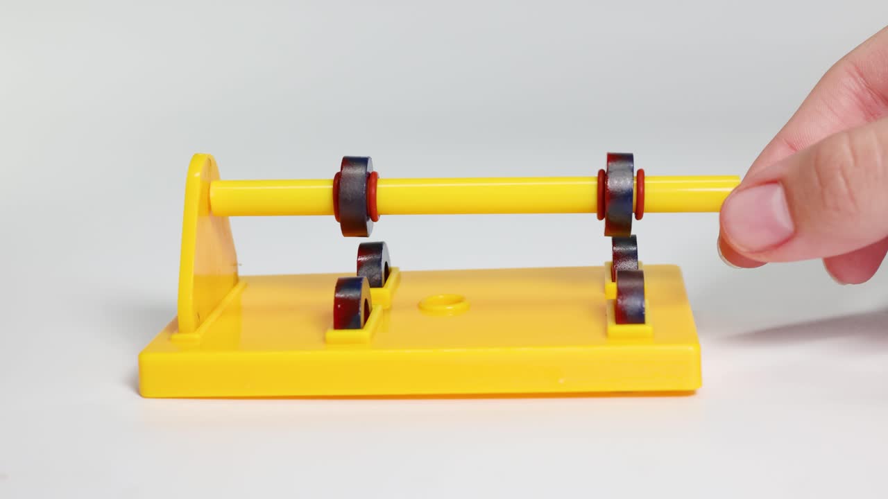 A hand manipulates a rod with magnetic rings, demonstrating attraction and repulsion on a yellow stand