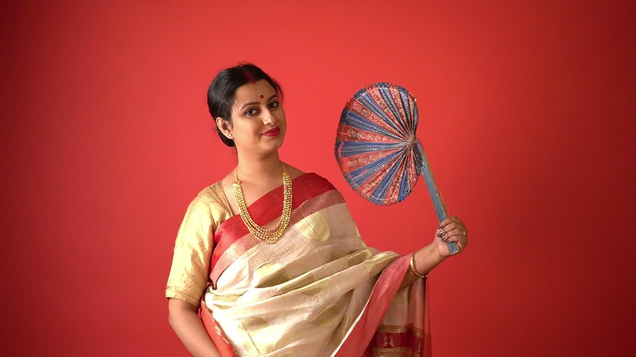 Kolkata : Traditionally dressed Bengali woman in Lal pad saree with gold jewelry standing against red background with beautiful handmade fan