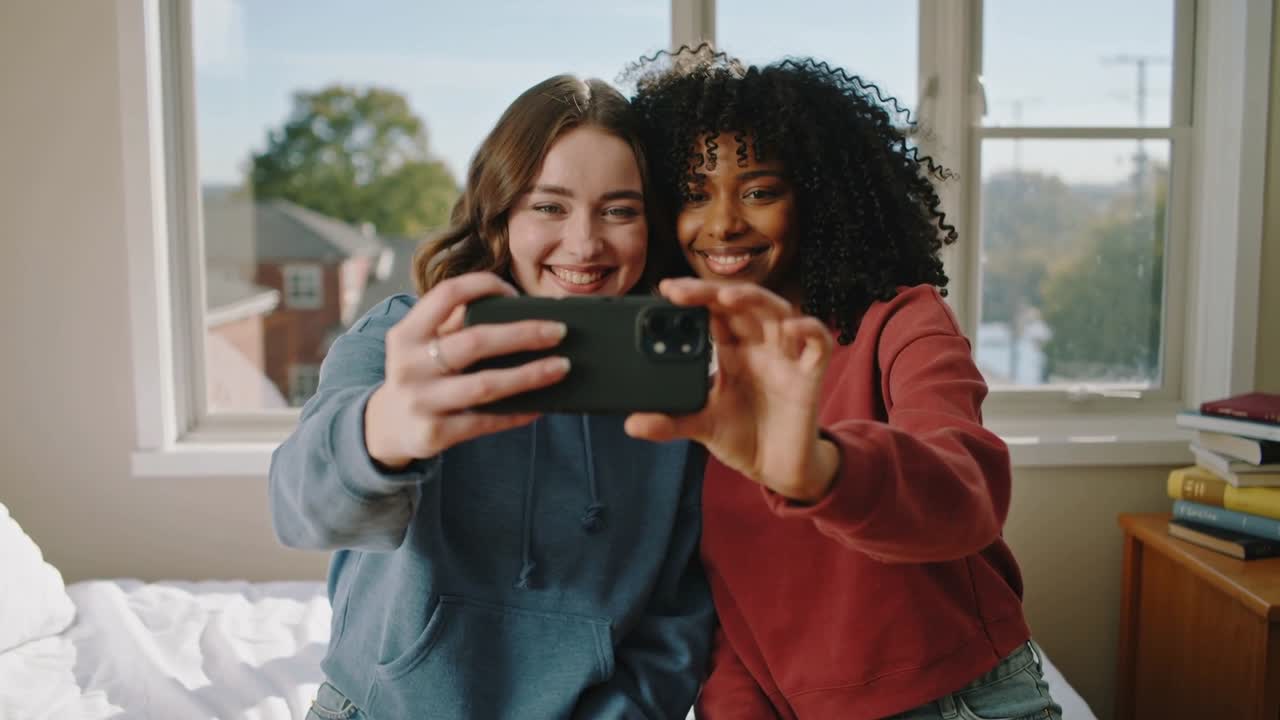 Donna con felpa blu con cappuccio che allunga il telefono per un selfie stabilizzandosi, mentre un'amica con felpa rossa sorride sul letto.