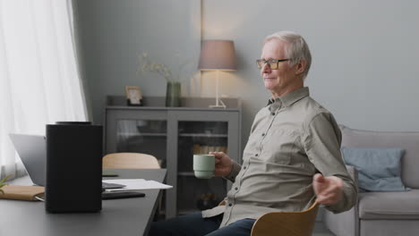 Happy-Elderly-Man-Listening-To-Music-While-He-Is-Sitting-At-Desk-Holding-A-Coffee-Cup