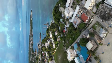 Drone-portrait-view-in-Belize-flying-over-caribbean-dark-and-light-blue-sea,-a-white-sand-caye-covered-with-palm-trees-and-restaurants-on-a-cloudy-day