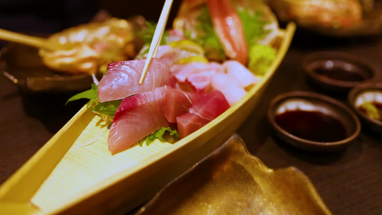 Primo piano di bacchette che sollevano pesce crudo da un tradizionale piatto giapponese a forma di barca per sashimi.