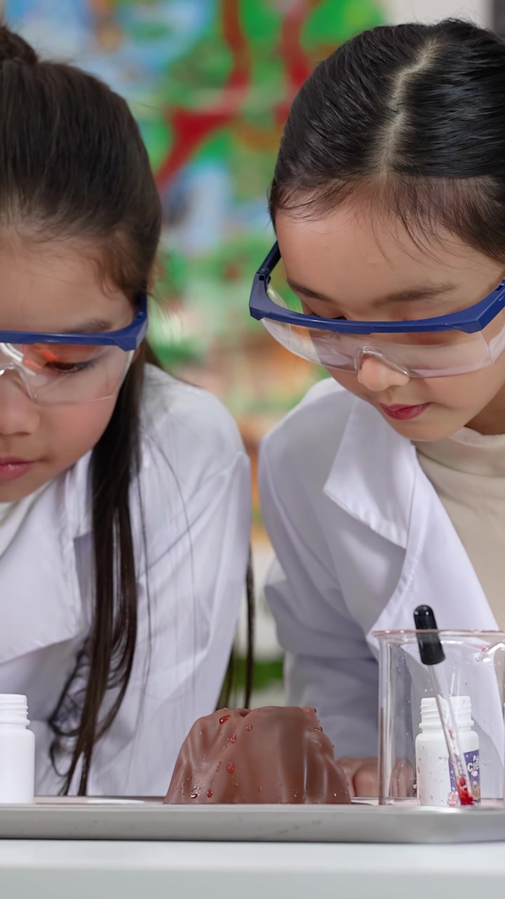 Dua gadis muda mengenakan kacamata pengaman dan jas lab melakukan percobaan sains gunung berapi.