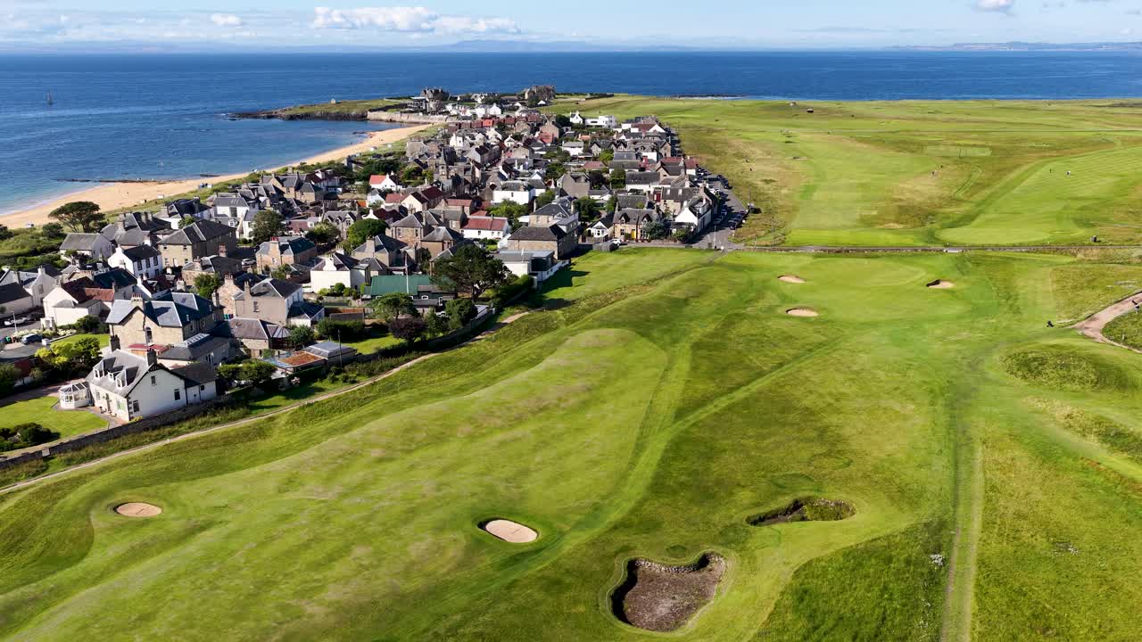 Drone glides above green golf course, seaside homes, and coastline under bright daylight, smooth movement