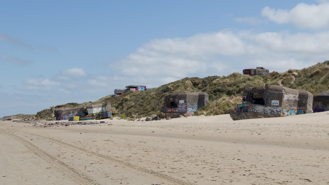 Wide shot of colorful graffiti-covered bunkers on sandy beach dunes under bright daylight, static camera