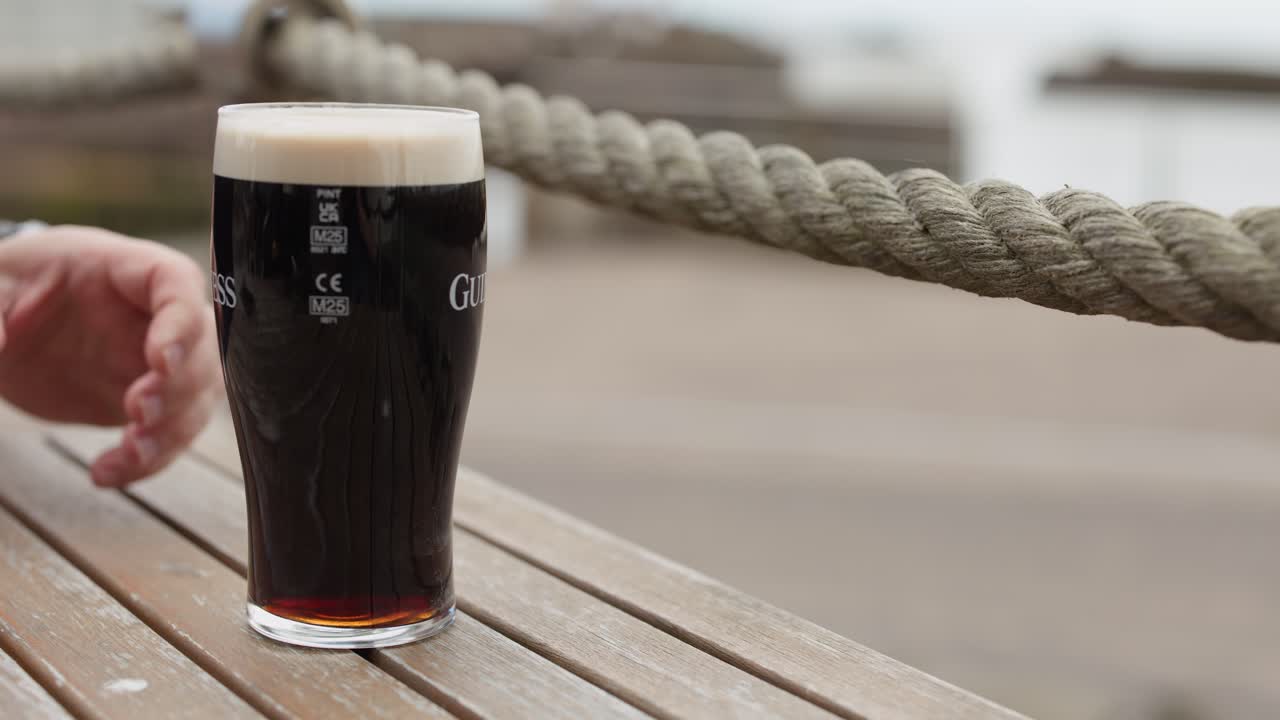Person lifts and sets down stout beer glass on wooden table, outdoor setting, natural light