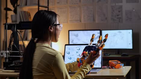 back view of teen asian girl with 3d printing assembling a cyborg hand while designing it on a laptop at home