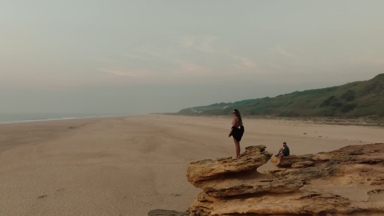 Aerial shot of a woman standing on a rocky ledge at sunset, gazing out at the ocean and empty sandy beach