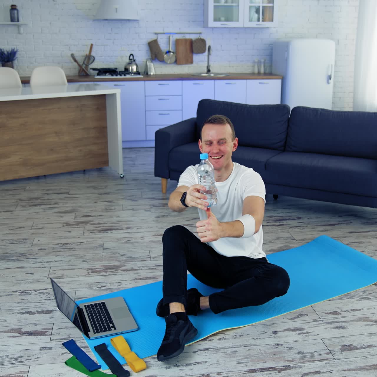 Young man exercising at home. Man is sitting on a mat on floor and holding balance with a bottle. Fitness at home during quarantine