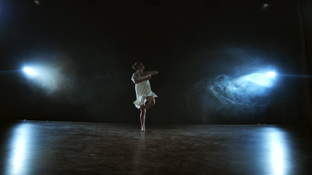 a young girl in a white dress dances a modern ballet makes rotations and jumps in slow motion on the stage with smoke in spotlights in full shot. Camera is zooming out standing behide the girl.