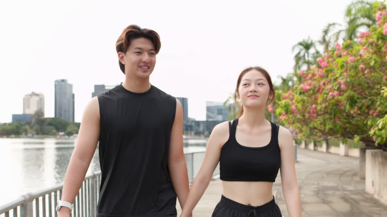 คู่รักนักกีฬาหนุ่มสาวเดินจูงมือกันไปตามทางเดินริมทะเลสาบในสวนสาธารณะในเมืองที่มีแดดส่อง