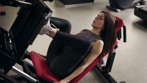atractiva y deportiva gitla morena de unos 20 años trabajando en una máquina de presión de piernas en el gimnasio. cámara lenta, en interiores.