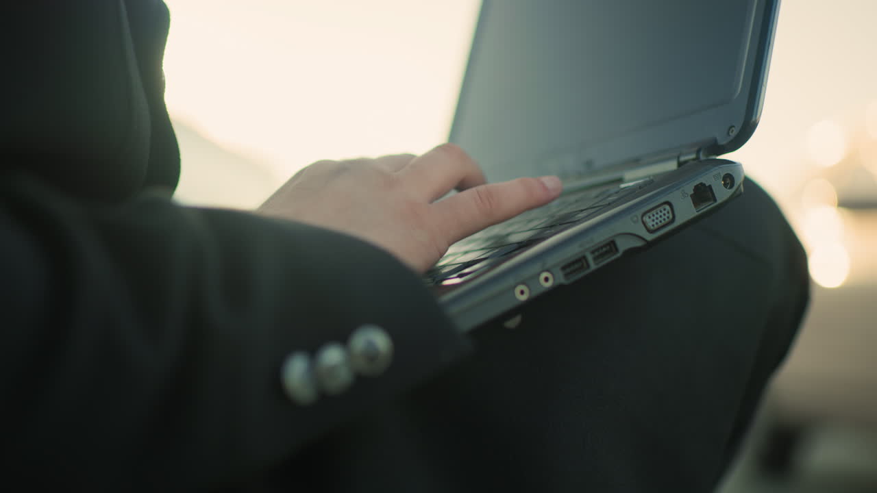 흑색 의복을 입은 개인의 손이 야외에서 노트북을 작동하고 초점과 정확성을 강조합니다. 세부 사항에는 의복 버튼, 노트북 포트 및 부드러운 보케 배경이 포함됩니다.