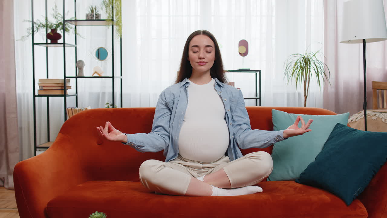 평온 한 임신 한 미소 짓는 여자 는 집 방 의 편안 한 소파 에서 명상 하는 동안 로터스 자세 에 앉아 있다