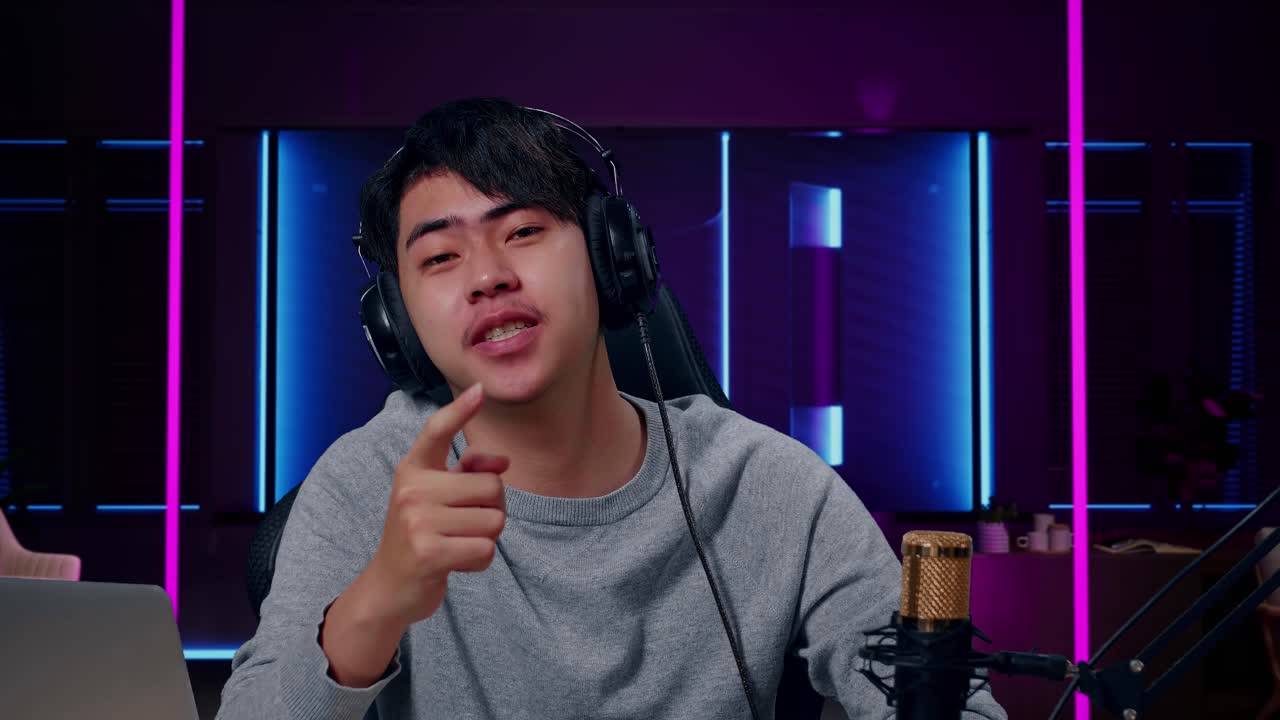 primer plano de un jugador asiático con auriculares y computadora agitando la mano y luego levantando el dedo índice mientras juega en una habitación iluminada por luces de neón en retro