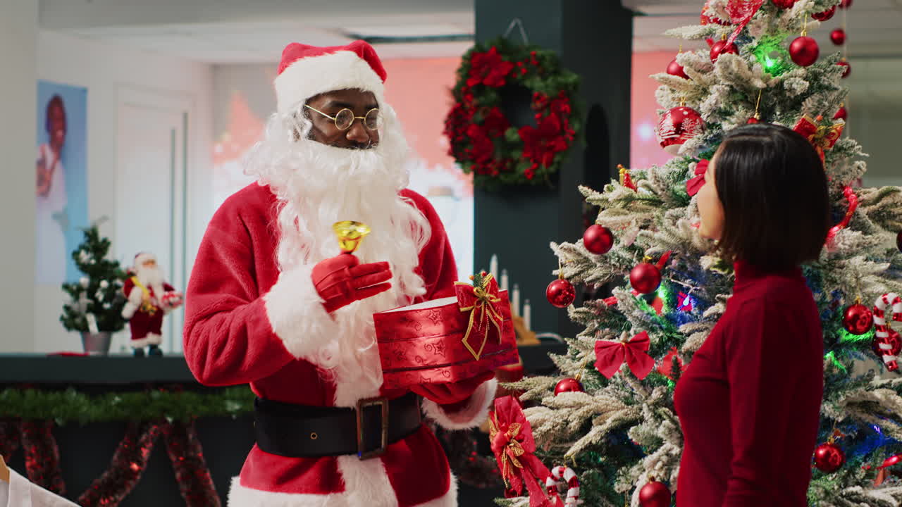 empleado afroamericano vestido de santa claus tocando la campana y sosteniendo el regalo de navidad junto al árbol adornado de navidad, hablando con un cliente asiático comprando en una tienda de moda decorada festiva