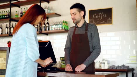 attractive young woman is talking to friendly male cashier, paying for takeaway coffee with mobile phone at coffee shop, then taking glass and leaving.