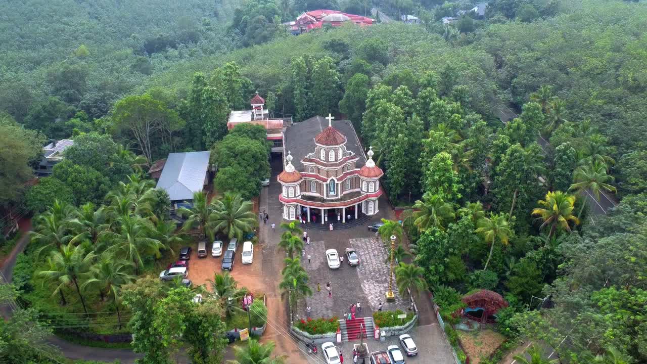 Wide aerial view of a beautiful, traditional Christian church with a distinctive facade and courtyard, entirely surrounded by dense tropical forest rubber plantation in Kerala, India