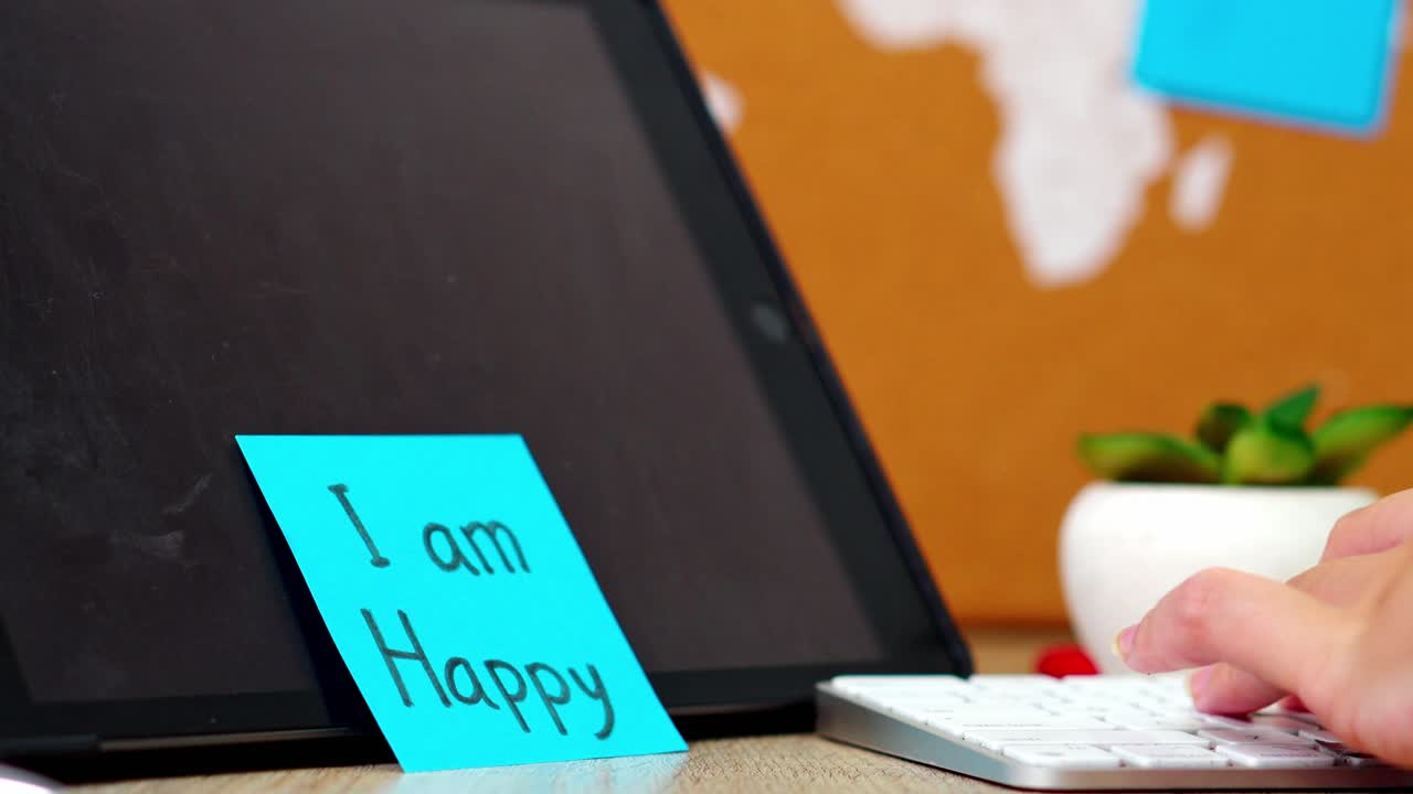 Close-up of Hand sticking blue "I am Happy" note on tablet screen while working at desk