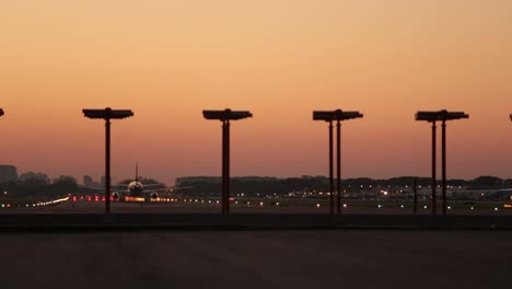 airplane taking off at dusk with runway lights and city silhouette in the background, warm sky