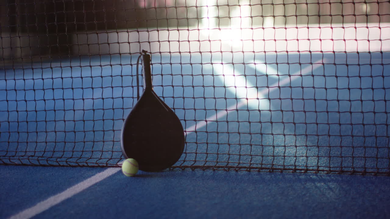 Padel racket and ball resting against net on blue court, copy space