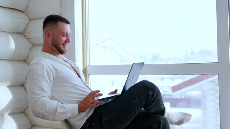happy laughing caucasian male in white shirt sits on the chair near the window. man holds a laptop and reads from it out loud. work online.